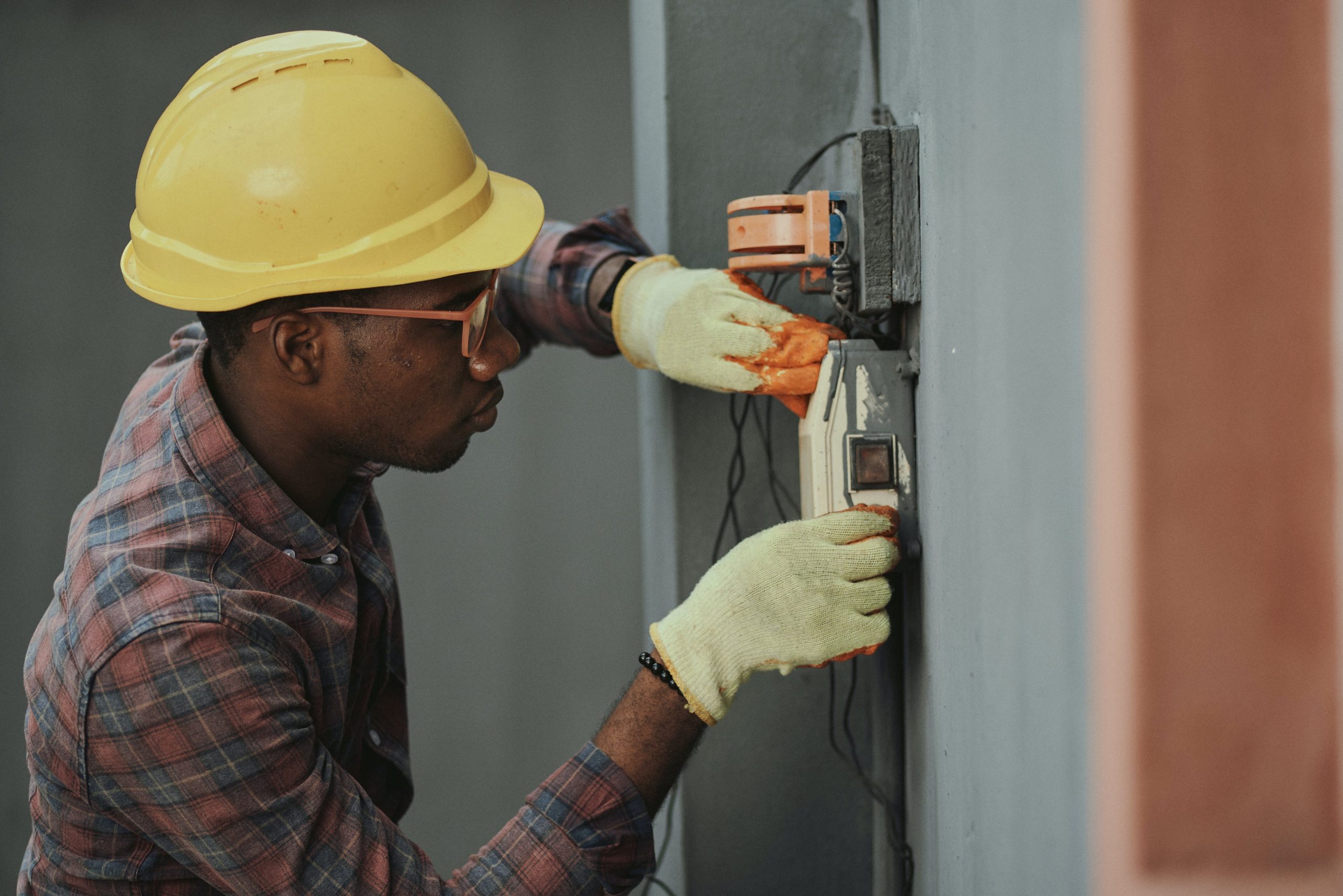 Man in Yellow Hat and Yellow Gloves Repairing a Fuse Box Attached to a Wall