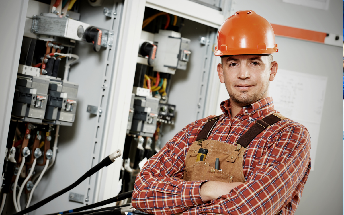 An Electrician Wearing a Hard Hat Crossing His Arms in Front of Electrical Equipment.