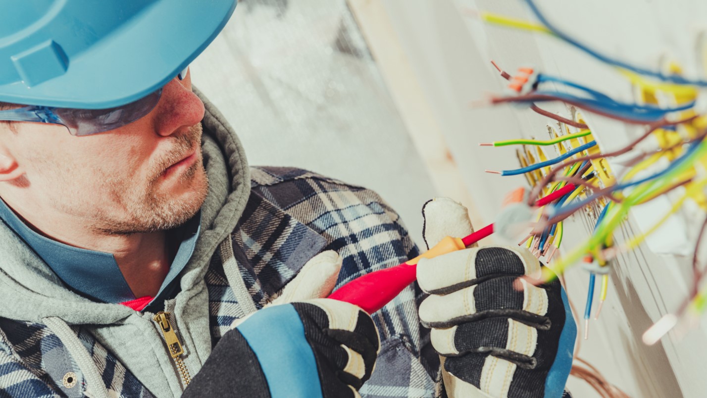 A close-up of an electrician in black gloves using a tool to tighten an electrical component.