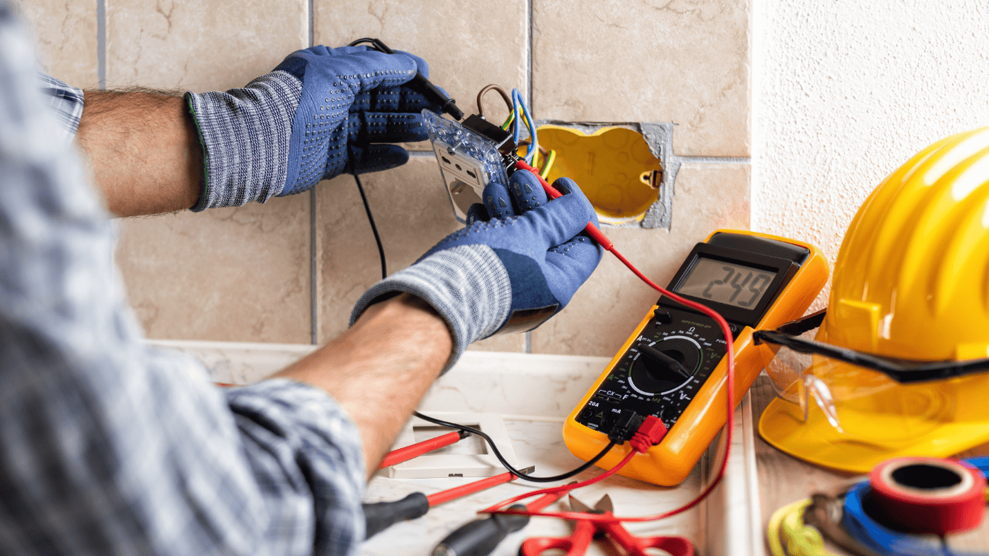 An electrician uses a tool to measure electricity, near the tool is a hard hat and safety glasses.